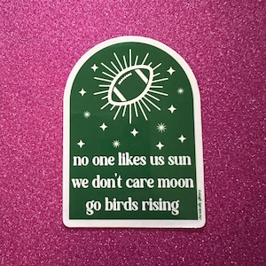 May include: Green sticker with a white border featuring a white football inside a sun with white rays. The text reads "no one likes us sun we don't care moon go birds rising".
