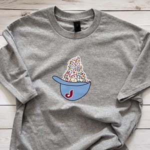 May include: A gray t-shirt featuring a blue baseball cap graphic filled with colorful sprinkles. The cap has a red and white "J" logo. The shirt is made of a soft material and has long sleeves.