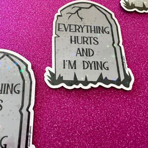 May include: A black and white sticker with a tombstone design. The tombstone has the text "Everything Hurts and I'm Dying" on it. The sticker is on a pink glitter background.