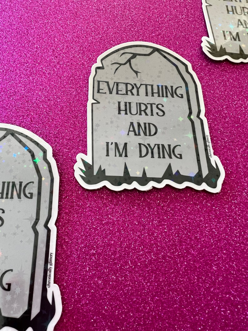 May include: A black and white sticker with a tombstone design. The tombstone has the text "Everything Hurts and I'm Dying" on it. The sticker is on a pink glitter background.