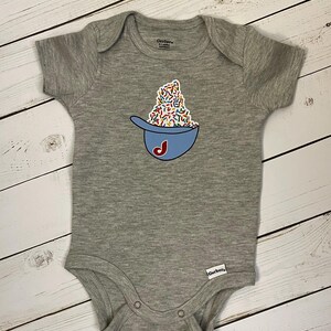 May include: A gray baby onesie with a blue baseball cap design filled with colorful sprinkles and a red "J" on the front. The onesie has short sleeves and snap closures.