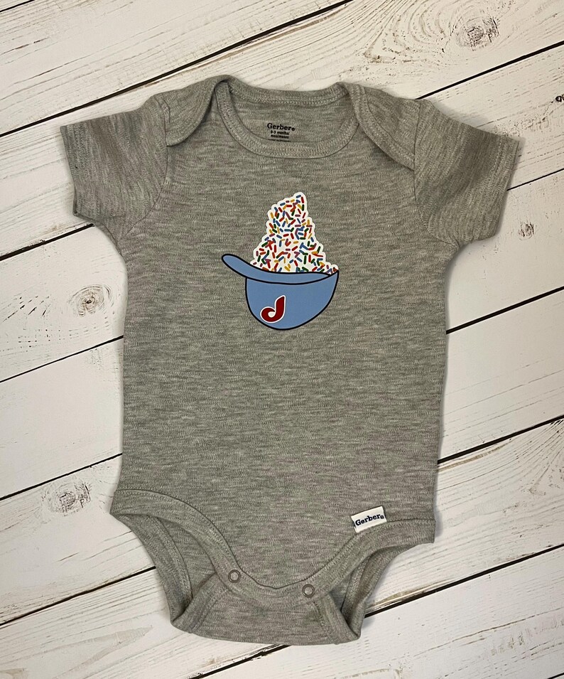 May include: A gray baby onesie with a blue baseball cap design filled with colorful sprinkles and a red "J" on the front. The onesie has short sleeves and snap closures.