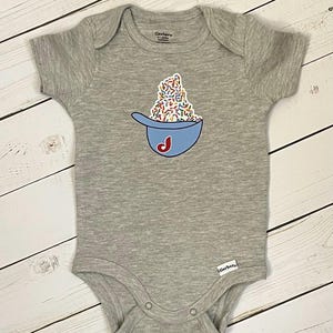 May include: A gray baby onesie with a blue baseball cap graphic filled with colorful sprinkles and a red "J" on the side. The onesie has short sleeves and snap closures. The brand name "Gerber" is visible.