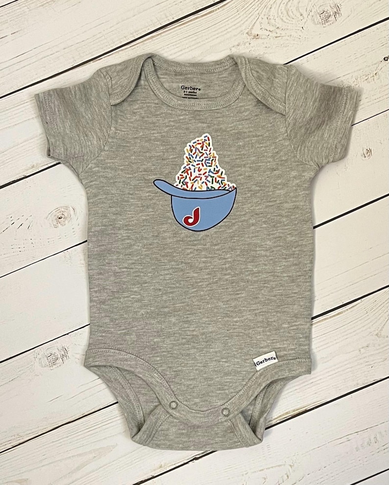 May include: A gray baby onesie with a blue baseball cap graphic filled with colorful sprinkles and a red "J" on the side. The onesie has short sleeves and snap closures. The brand name "Gerber" is visible.