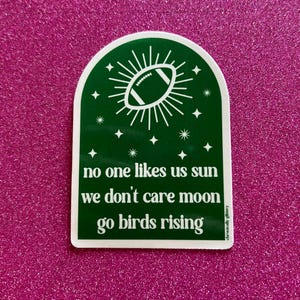May include: Green and white sticker with a football shaped sun and stars. The text reads "no one likes us sun we don't care moon go birds rising".