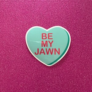 May include: A light blue heart-shaped sticker with the text "BE MY JAWN" in red letters. The sticker is on a pink glitter background.