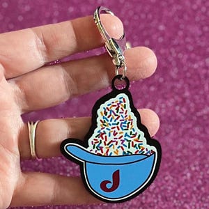 May include: A blue bowl-shaped keychain with a black outline, filled with colorful sprinkles. The bowl has a red "J" on it. The keychain is attached to a silver clasp.