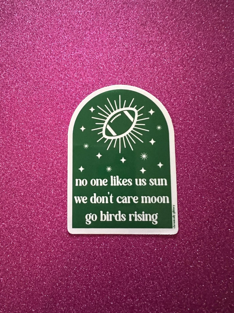 May include: Green sticker with a white border featuring a white football inside a sun with white rays. The text reads "no one likes us sun we don't care moon go birds rising".