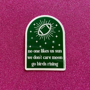 May include: Green and white sticker with a football and stars. The text reads "no one likes us sun we don't care moon go birds rising".