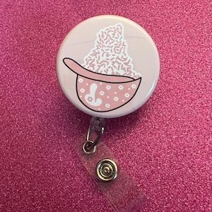 May include: A light pink badge reel with a cartoon illustration of a pink bowl filled with pink sprinkles. The bowl has a handle and is decorated with white flowers and a stylized design. A clear plastic strap and metal clip are attached.