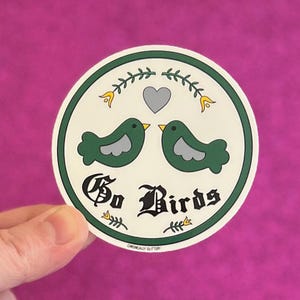 May include: A round sticker with a green border and the text "Go Birds" in a gothic font. The sticker features two green birds facing each other, with a gray heart above them. The background is a vibrant pink.