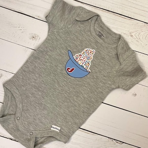 May include: A gray baby onesie with a blue baseball cap graphic filled with colorful sprinkles. The onesie has short sleeves and snap closures at the bottom. The Gerber brand label is visible.