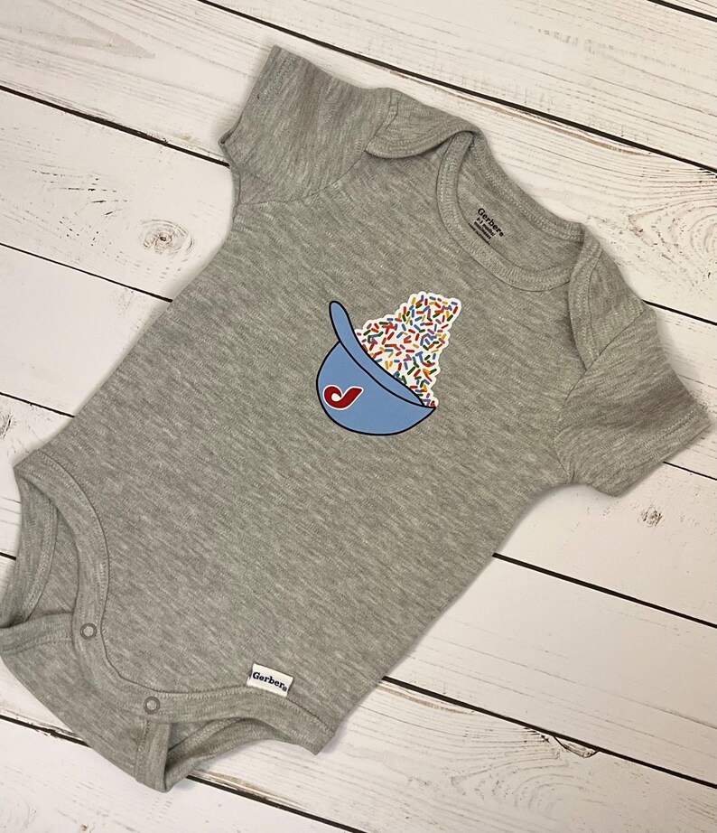 May include: A gray baby onesie with a blue baseball cap graphic filled with colorful sprinkles. The onesie has short sleeves and snap closures at the bottom. The Gerber brand label is visible.
