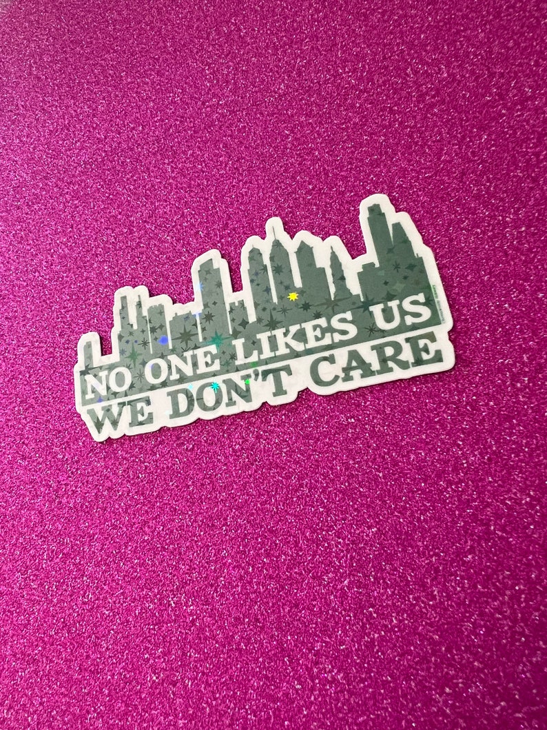 May include: A white sticker with a black and gray cityscape silhouette and the text "NO ONE LIKES US WE DON'T CARE" in black.