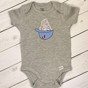 May include: A gray baby onesie with a blue baseball cap design filled with colorful sprinkles and a red "J" initial. The onesie has short sleeves and snap closures. The Gerber brand label is visible.