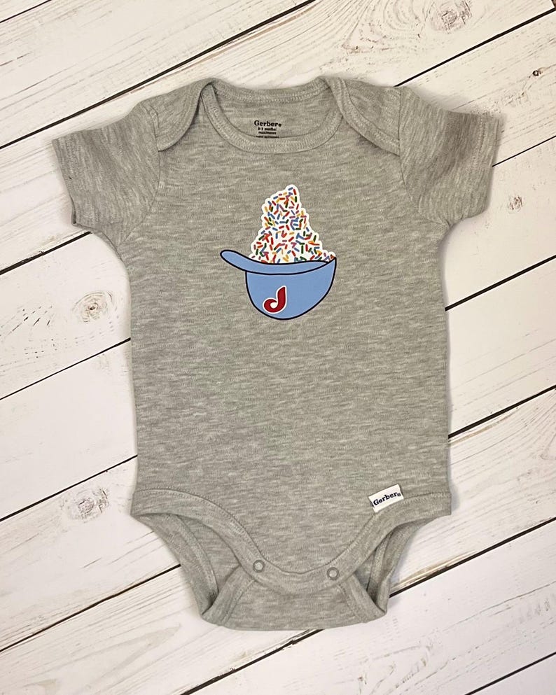 May include: A gray baby onesie with a blue baseball cap design filled with colorful sprinkles and a red "J" initial. The onesie has short sleeves and snap closures. The Gerber brand label is visible.