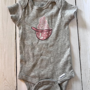 May include: A gray baby onesie with a pink and white cupcake design. The cupcake is in a pink bowl with white flower accents and a pink handle. The onesie has short sleeves and a snap closure.