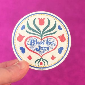 May include: A round sticker with a floral design and the text "Bless this Jawn" in a heart shape. The sticker features red, blue, and green accents, with a dotted border and a glittery finish. The sticker is held by a hand against a magenta background.