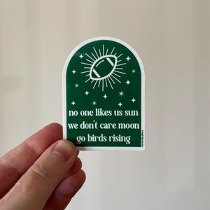 May include: Green sticker with a white football in a sunburst design. The text reads "no one likes us sun we don't care moon go birds rising".