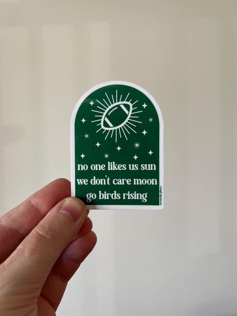 May include: Green sticker with a white football in a sunburst design. The text reads "no one likes us sun we don't care moon go birds rising".