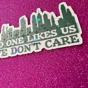 May include: A white sticker with a black silhouette of a city skyline and the text "No one likes us, we don't care". The sticker has a glittery finish.