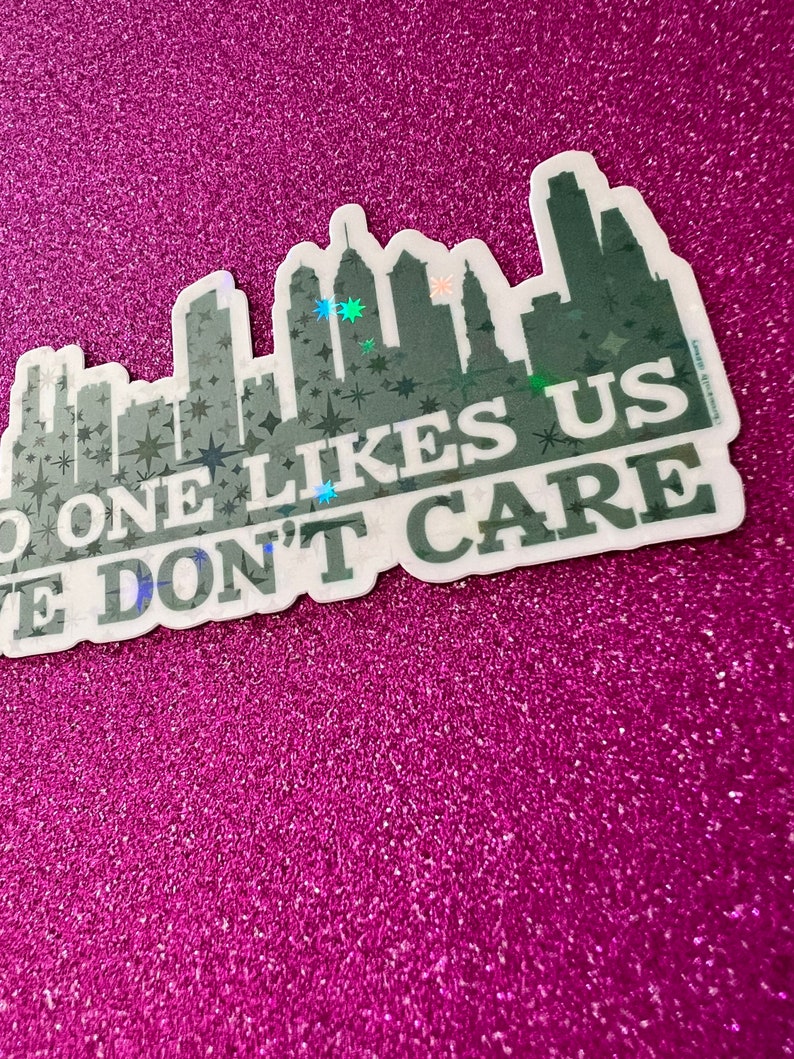 May include: A white sticker with a black silhouette of a city skyline and the text "No one likes us, we don't care". The sticker has a glittery finish.