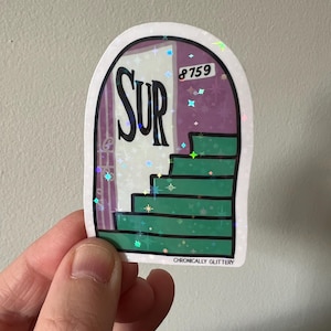 May include: A holographic sticker featuring a purple door with the word "SUR" on it and a green staircase leading up to the door. The door has the number "8759" on it. The sticker has a glittery finish and the text "CHRONICALLY GLITTERY" at the bottom.