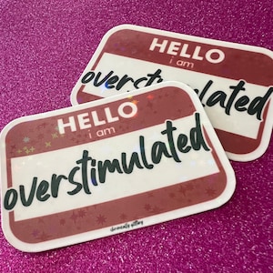 May include: Two brown name tag stickers with white text that reads "Hello I am Overstimulated". The stickers have a glittery finish and a white background.