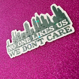 May include: A white sticker with a black cityscape silhouette and the text "No one likes us we don't care". The sticker has a holographic finish.