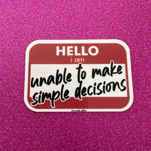 May include: A red and white name tag sticker with the text "HELLO i am unable to make simple decisions" in black cursive font.