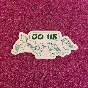 May include: A white sticker with four cartoon birds and the text "GO US" in a green speech bubble. The birds are outlined in green and are on a pink glitter background.