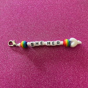 May include: White beaded keychain with a rainbow charm and the words "SHE HER" in black letters.