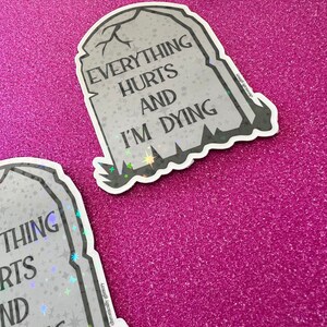 May include: A gray tombstone sticker with the text "Everything Hurts And I'm Dying" in black lettering. The sticker has a glittery finish and a black outline.