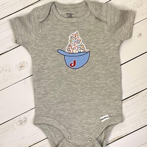 May include: A gray baby onesie with a blue bowl graphic filled with colorful sprinkles and a red "J" on the side. The onesie is made of soft material and has short sleeves. The Gerber brand label is visible.
