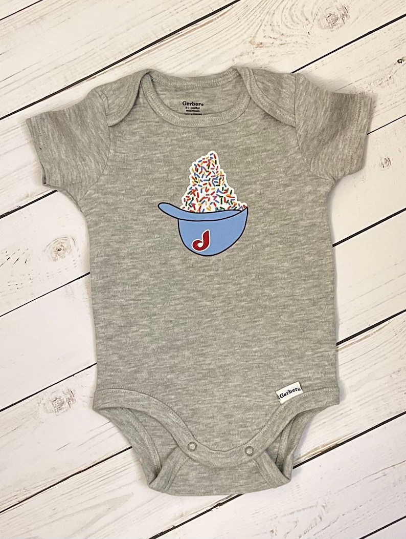 May include: A gray baby onesie with a blue bowl graphic filled with colorful sprinkles and a red "J" on the side. The onesie is made of soft material and has short sleeves. The Gerber brand label is visible.