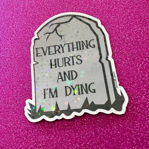 May include: A white and gray holographic sticker with a tombstone shape. The sticker has the text "Everything Hurts And I'm Dying" on it.