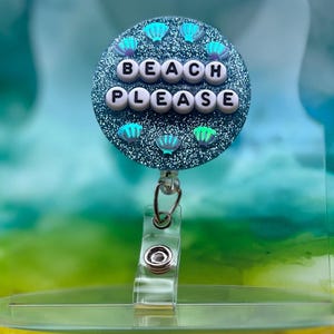 May include: A blue glitter badge holder with a white background and the words "Beach Please" in white letters. The badge holder has a clear plastic clip.