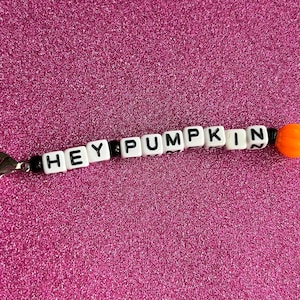 May include: A white and black beaded keychain with the words "HEY PUMPKIN" spelled out in black letters on white beads. The keychain also features a small orange pumpkin bead.