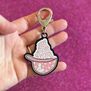 May include: A pink and white keychain featuring a bowl of what appears to be ice cream or a dessert, with pink sprinkles. The bowl has a black outline and a handle. The keychain has a silver clasp.