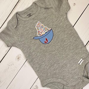 May include: A gray baby onesie with a blue baseball cap design filled with colorful sprinkles and a red "J" logo. The onesie is made of soft material and has short sleeves. The Gerber brand label is visible.