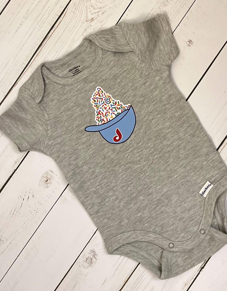 May include: A gray baby onesie with a blue baseball cap design filled with colorful sprinkles and a red "J" logo. The onesie is made of soft material and has short sleeves. The Gerber brand label is visible.