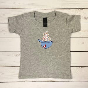 May include: A gray toddler t-shirt featuring a blue baseball cap graphic filled with colorful sprinkles. The cap has a red "J" on it. The shirt is made of a soft material and has a crew neck.