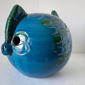 Könnte beinhalten: Eine tealblaue Keramik-Sparbüchse mit grünem Kamm und einer strukturierten Oberfläche. Die Sparbüchse hat ein großes Auge und eine kleine Schnauze.