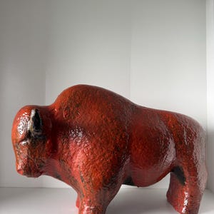 Può includere: Scultura in ceramica di un bisonte in posizione eretta. Il bisonte è principalmente di colore rosso-arancio strutturato con dettagli neri su zampe e testa. Esposto su una mensola bianca.