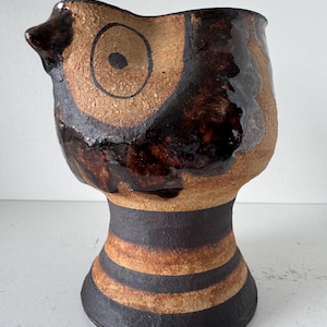 May include: A ceramic pitcher with a textured tan surface and dark brown glazed accents. The upper portion features a stylised eye design, while the base is decorated with horizontal dark brown stripes. The vessel has a wide rim and a small spout.