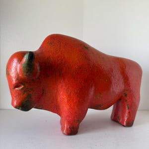 Può includere: Una scultura di bisonte in ceramica rossa con superficie strutturata e dettagli verde scuro. Il bisonte è raffigurato in posizione eretta, mostrando la sua forma robusta e i dettagli. È un oggetto decorativo.