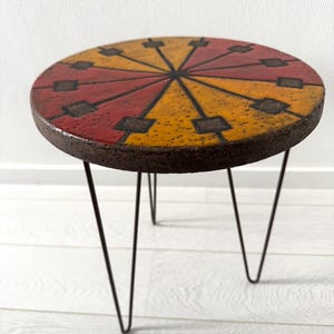 Op de afbeelding: Een kleine, ronde bijzettafel met een kleurrijk, geometrisch ontwerp op het tafelblad. De bovenkant heeft een patroon van rode, gele en bruine segmenten. De tafel heeft drie zwarte haarspeldpoten, wat een mid-century modern esthetiek creëert.