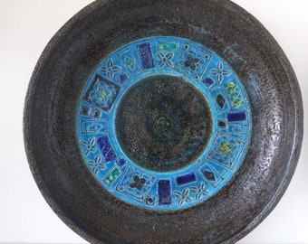 May include: A large, round ceramic plate with a dark brown glaze and a blue and green geometric design. The design features a circle in the center with a repeating pattern of squares, triangles, and circles.