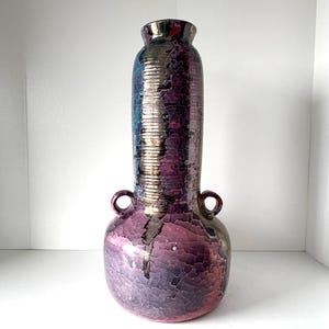 May include: A tall, decorative vase with a metallic, iridescent finish. The vase features a bulbous base, a long, cylindrical body with vertical ridges, and two small handles. The colour transitions from purple to blue and gold.