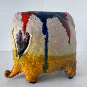 Puede incluir: Un jarrón de cerámica hecho a mano con un cuerpo beige texturizado. La parte superior presenta goteos de esmalte rojo y azul. La parte inferior está pintada de amarillo y naranja. El jarrón tiene tres pies pequeños y un asa curva.
